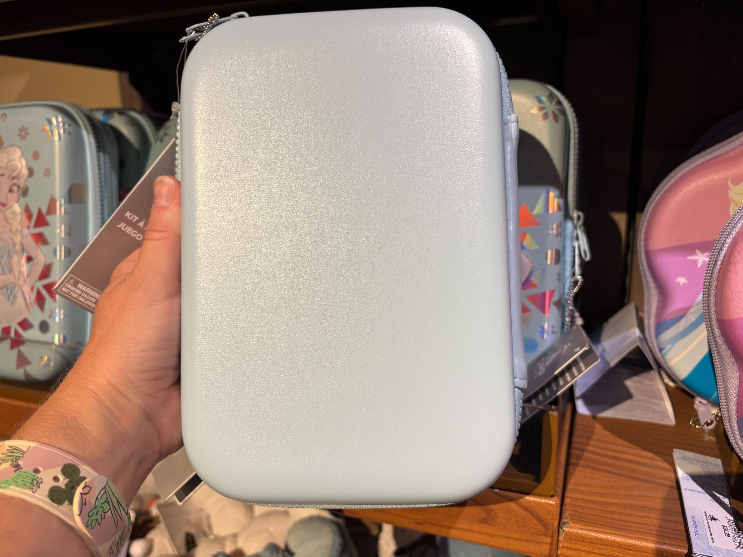 A hand holds a plain light blue rectangular case in a store, with Disney-themed bags on the shelf behind it.