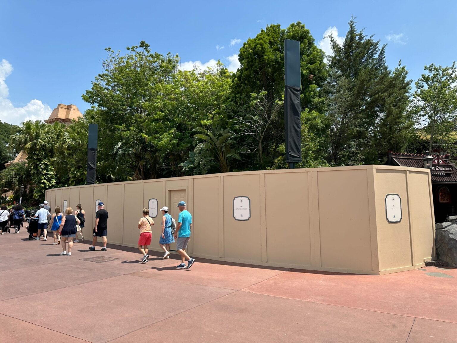 Construction walls curve around trees near a theme park walkway as people walk by under a partly cloudy sky.