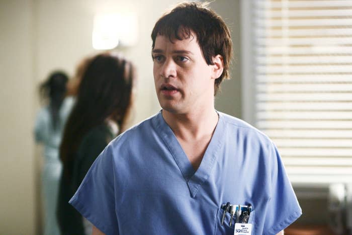 A person wearing scrubs stands in a medical setting, with a focused expression and pens clipped to the shirt pocket