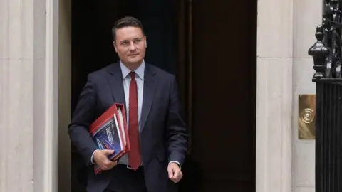 PA Media Health Secretary Wes Streeting, wearing a dark suit and red tie, leaves 10 Downing Street holding a red folder 