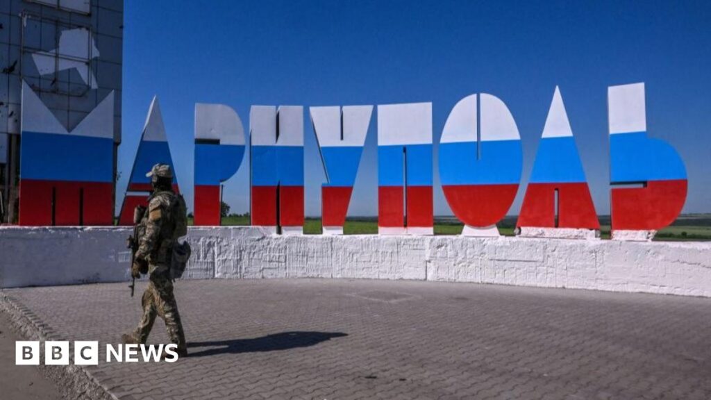 A Russian soldier stands in the ruins of Mariupol's theatre in spring 2022, taking a photo out of a destroyed window using his phone