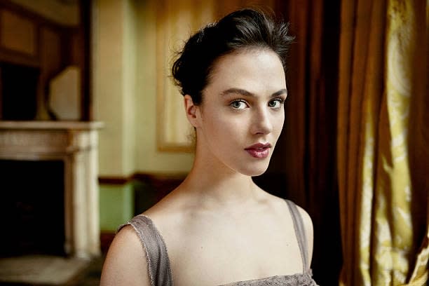 Person in elegant attire stands indoors, gazing directly at the camera, with softly lit background and draped curtains
