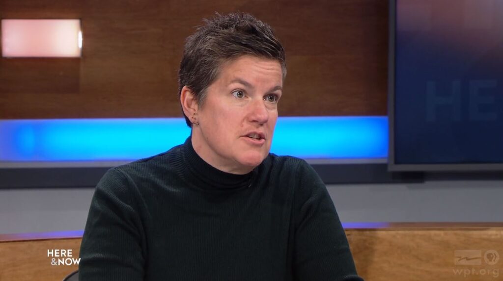 A person with short hair and a dark turtleneck speaks in a studio setting with a Here & Now logo visible on a screen and table.