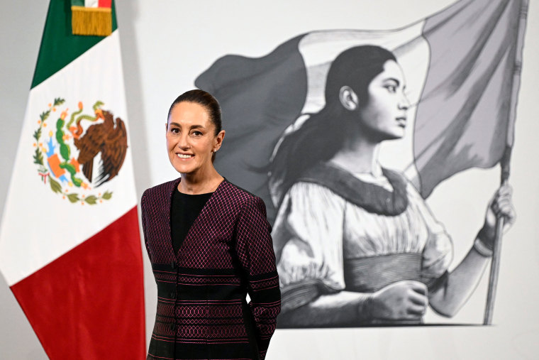 Claudia Sheinbaum  smiling in front a Mexican flag and mural