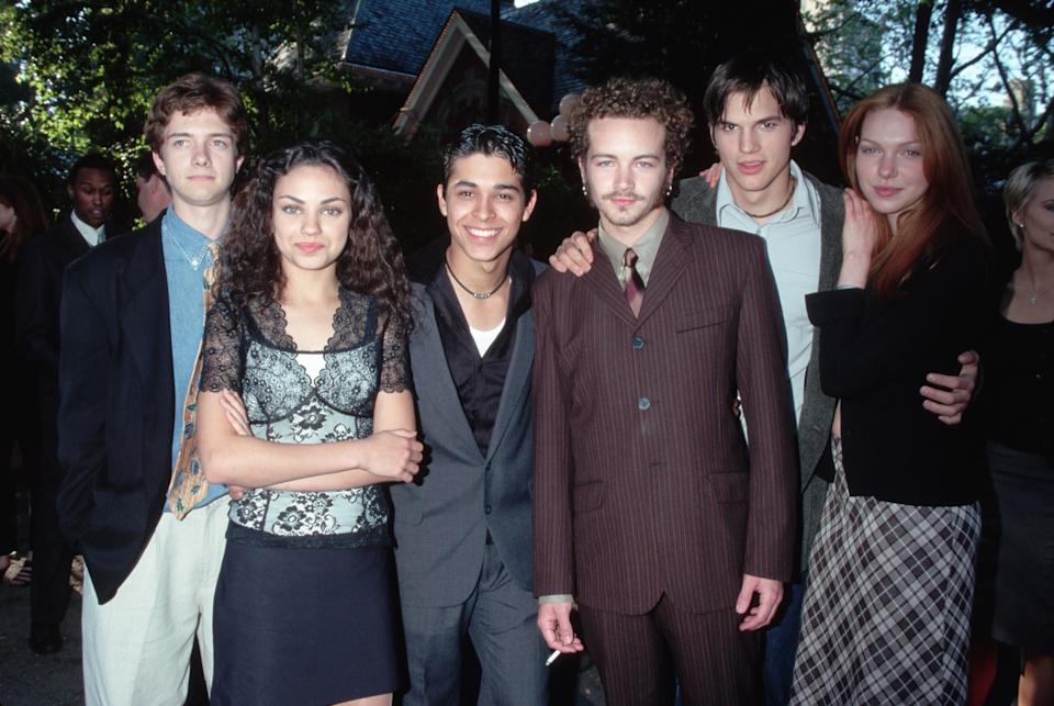 Group of six people posing together outdoors, with some dressed in 90s-style formal and casual attire