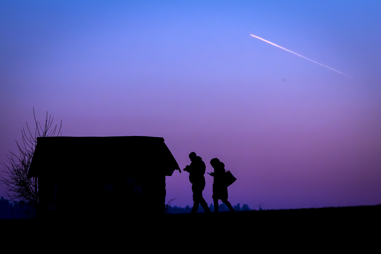 Two people in silhouette look at mobile phones while a contrail of an aircraft is seen in the sky in the evening twilight, at Zurich Kloten Airport, pictured in Ruemlang, Switzerland, Tuesday, Jan. 14, 2025. (KEYSTONE/Michael Buholzer)