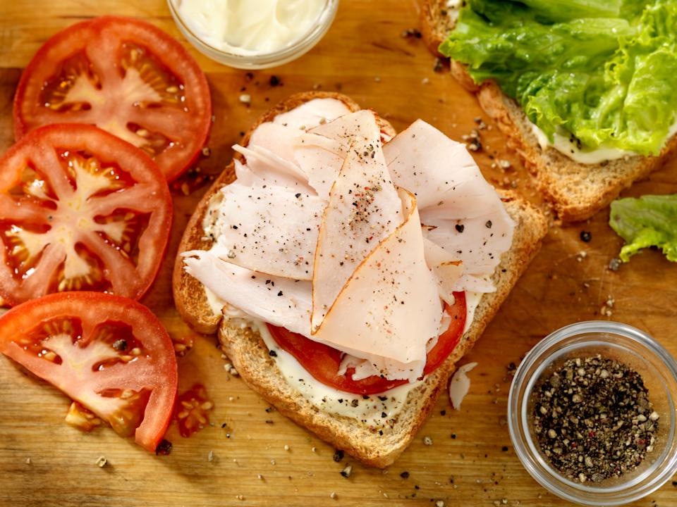 A turkey or chicken sandwich with tomatos and mayonnaise sits on a cutting board surrounded by ingredients.