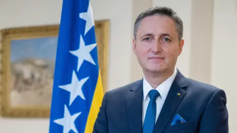 Bosnia and Herzegovina UK Network President of Bosnia-Herzegovina, Denis Bećirović, is wearing a navy blue suit jacket with a white shirt and blue tie, and is stood next to the Bosnia-Herzegovina national flag, which is blue and yellow with white stars on the blue section.