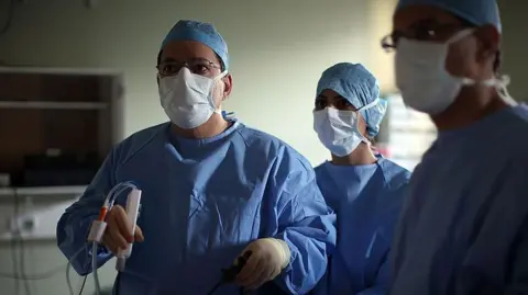 Getty Images A surgeon and his theatre team perform key hole surgery to remove a gallbladder at the Queen Elizabeth hospital in Birmingham. The doctors are all wearing blue overalls and white masks and staring off camera. The main surgeon is holding a medical device.