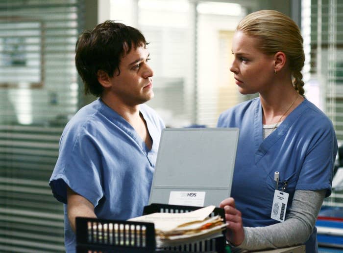 Two people in hospital scrubs have a serious conversation in a medical setting. One holds a folder with paperwork