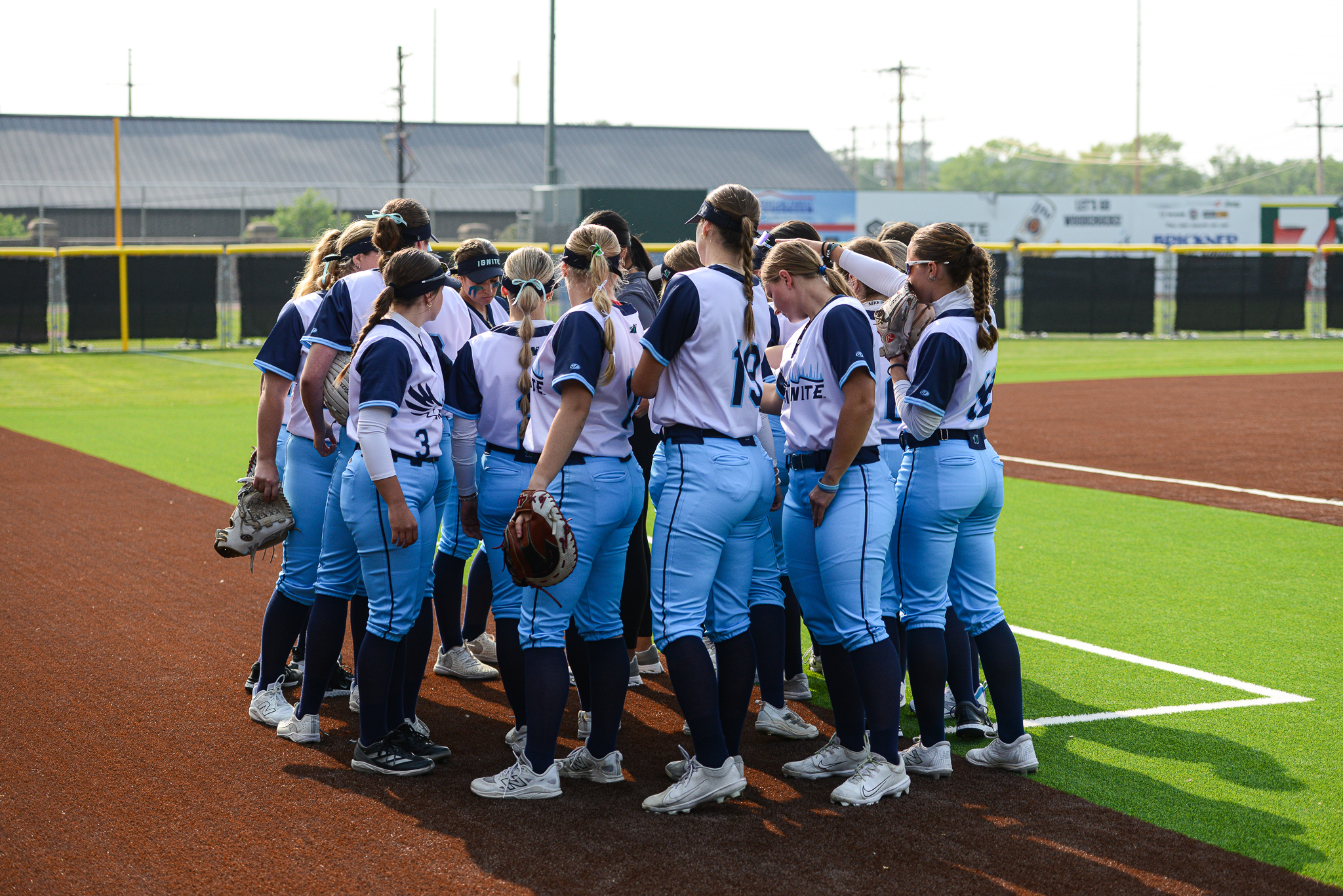 A softball team in blue uniforms stands together in a huddle on an outdoor field near the baseline.