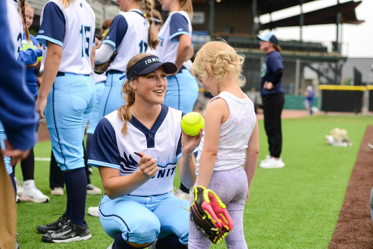 A softball player in uniform kneels and hands a softball to a young girl wearing a glove, with other players and people visible in the background on a field.