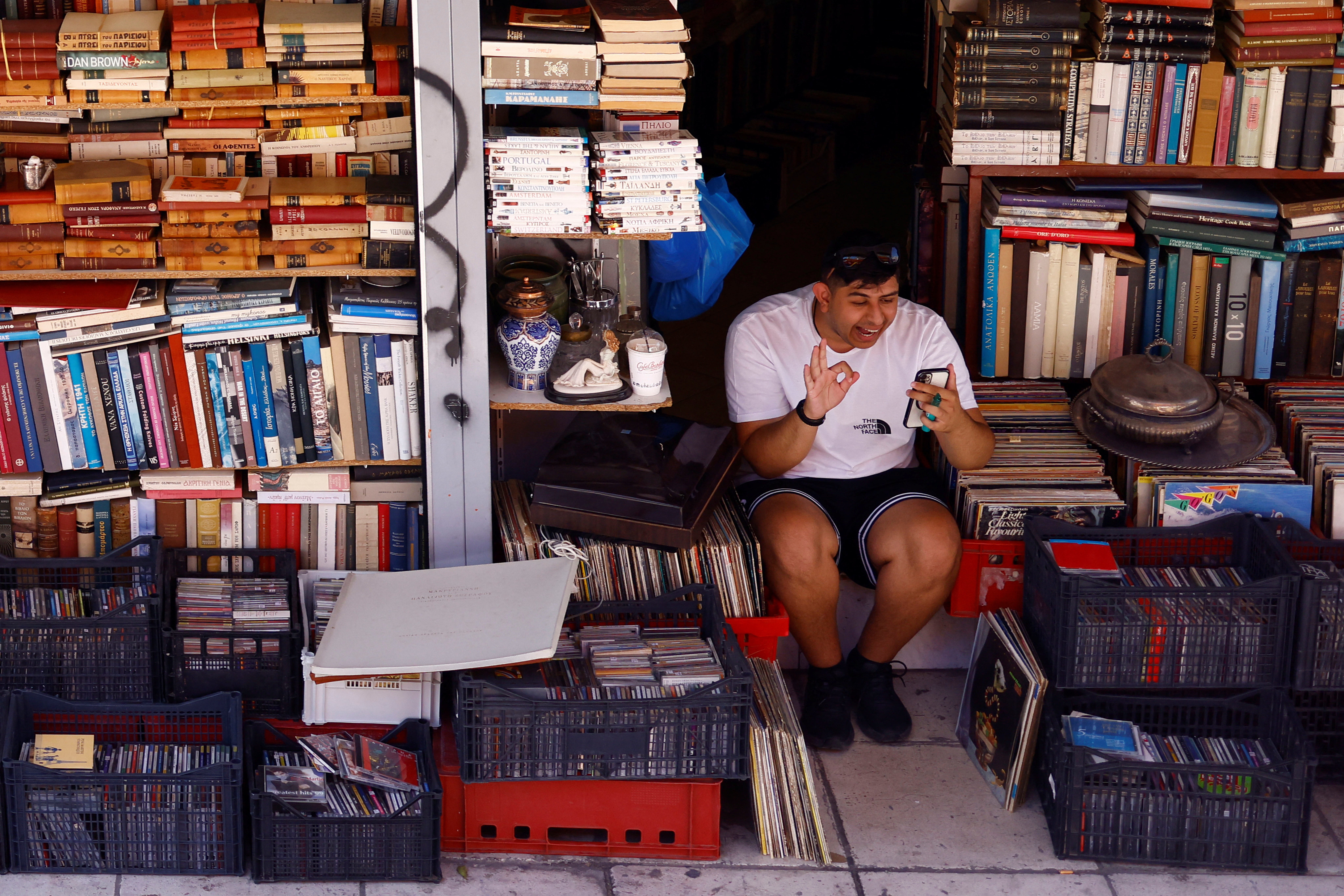A man gestures as he speaks on his mobile phone while taking shelter from the sun at a bookshop, as a heatwave hits Athens