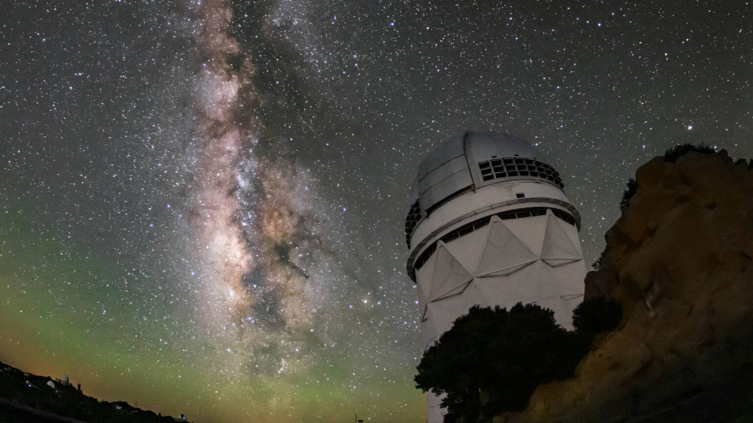 A white structure with a domed top on a rocky outcrop below a star-filled night sky. The Milky Way stretches vertically in a luminous band. Faint green glow tints the curved horizon.