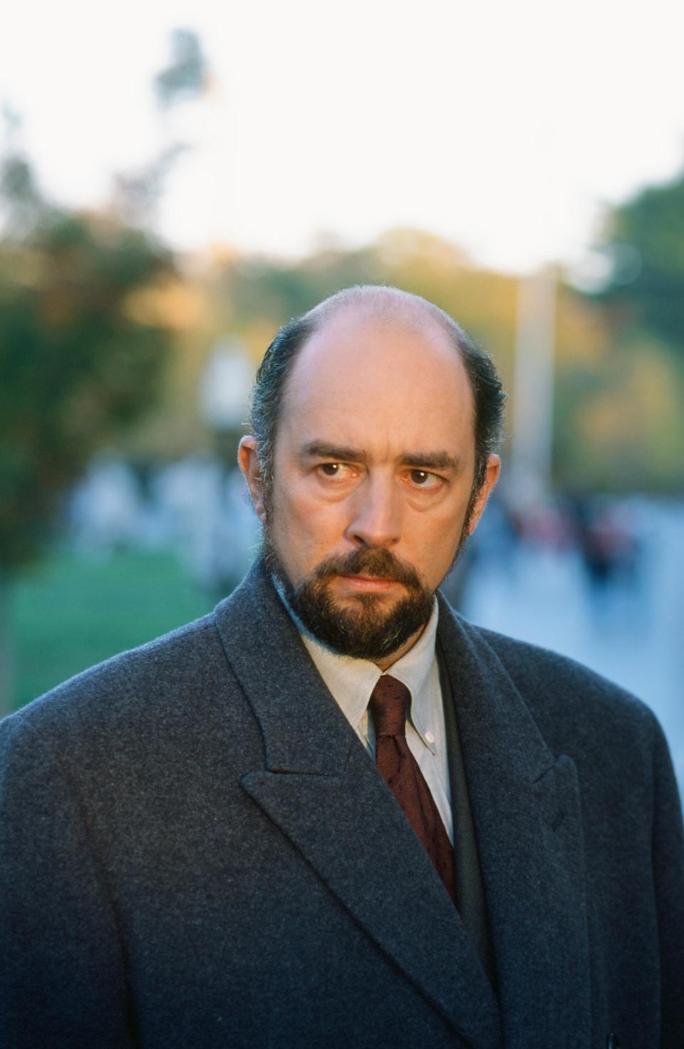 A man in a formal coat and tie stands outdoors with a serious expression