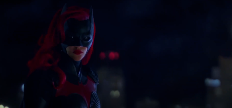 Person in dramatic, dark superhero costume with bat-like mask and cape, standing against a cityscape background at night