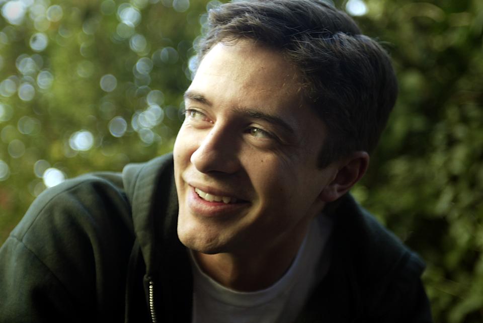 A smiling person wearing a casual hoodie outdoors, surrounded by blurred greenery in the background
