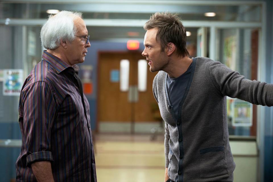 Two men in a hallway engage in a heated conversation; one wears a striped shirt, the other a cardigan over a t-shirt