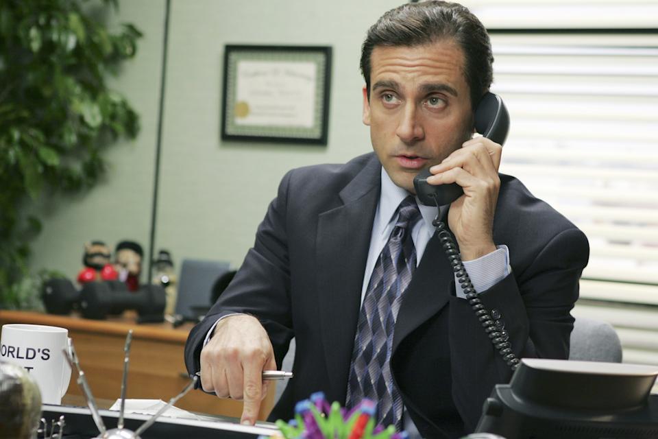 Man in a suit talks on the phone, seated at a desk with office decor. Holding a World's Best Boss mug, he looks focused