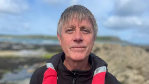 BBC A man looking directly at the camera, he is wearing a black zip up jacket with red and silver. Behind him the background is blurred. It is a rocky terrain and there is the sea in the background. 