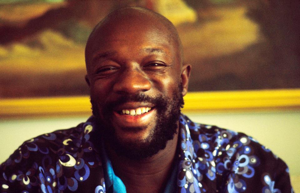 A smiling man with a beard, wearing a patterned shirt with circular designs, stands in front of a framed painting