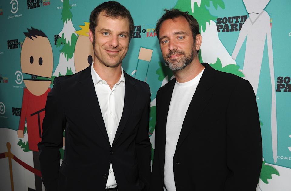 Two men in suits stand in front of a "South Park" backdrop featuring cartoon characters. They are smiling and posing for the camera
