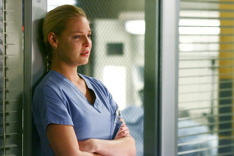 Person in medical scrubs stands with arms crossed, looking pensive in a clinical setting with a clipboard under one arm