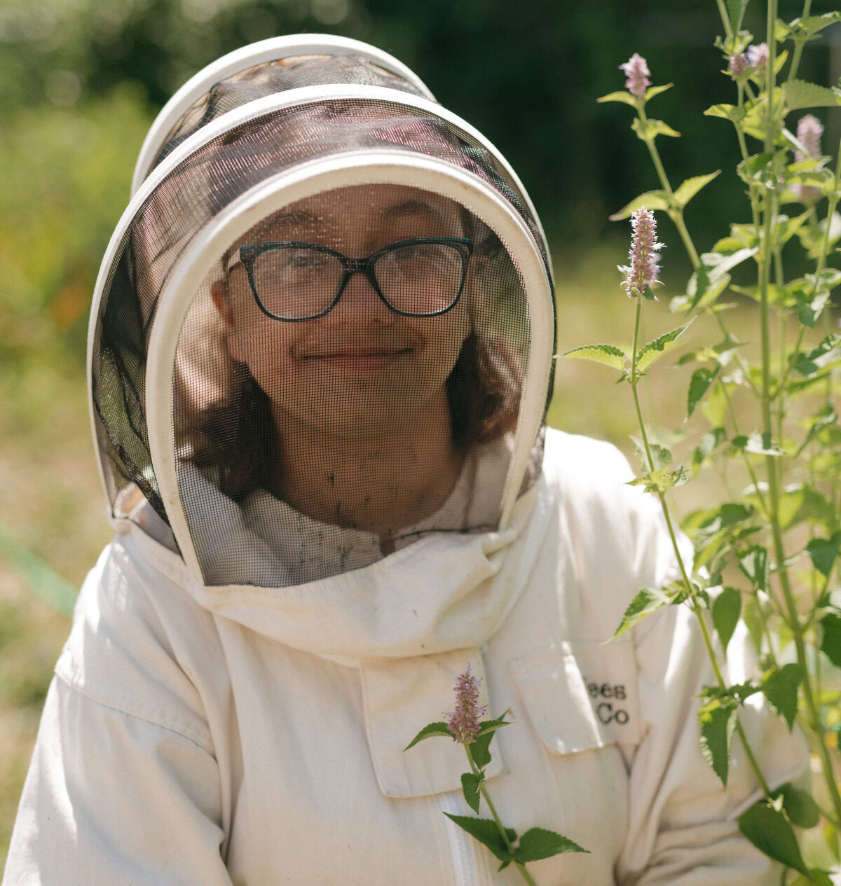 Alex Guzman is a junior beekeeping instructor with the Huneebee Project. She stands by some plants wearing a white beekeeper suit.