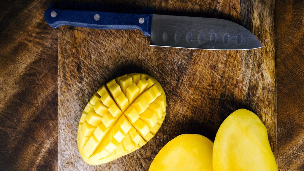 Mango halves diced on a chopping board with skin still intact as the knife lays next to them