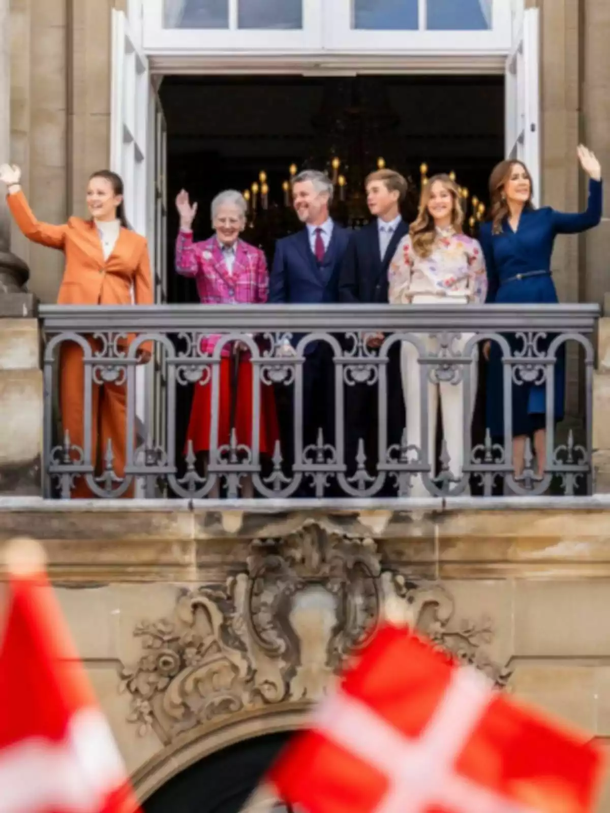 Six elegantly dressed people wave from a decorated balcony while several Danish flags flutter in the foreground.