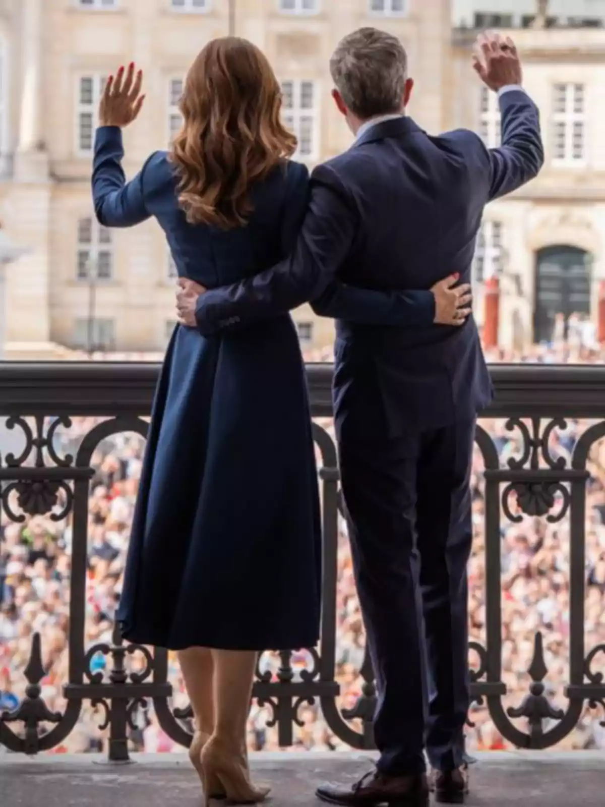Mary and Frederik of Denmark, elegantly dressed, wave to a crowd from a balcony.