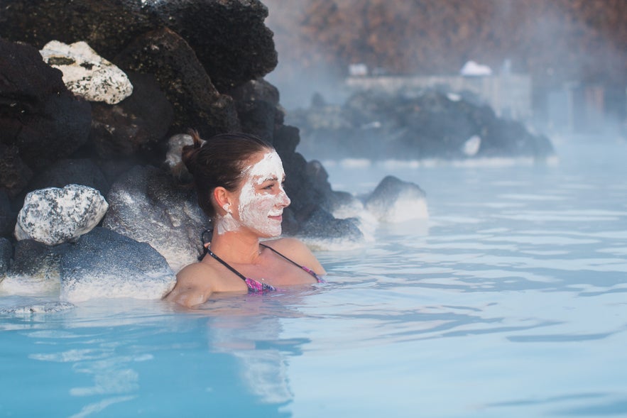 A smiling woman with a white mud mask enjoys the warm, milky-blue waters of Iceland's Blue Lagoon.