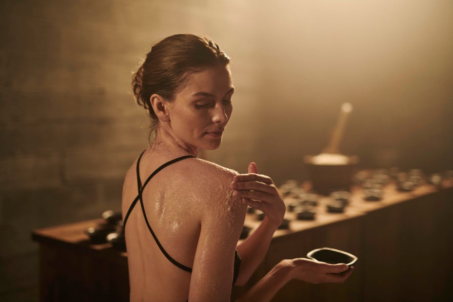 A person with hair tied back applies a body scrub to their shoulder at Sky Lagoon, holding a small bowl.