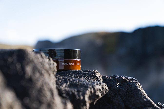 A small jar of Sky Lagoon Body Scrub sits on a dark volcanic rock, set against a blurred Icelandic landscape background.