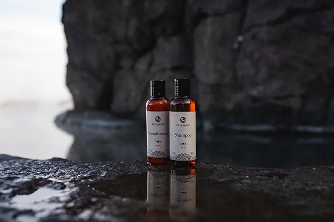 Sky Lagoon shampoo and conditioner bottles sit on dark, wet rocks by the water, with blurred cliffs in the background.