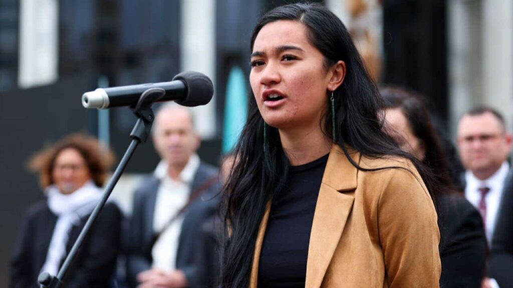 A woman in a beige blazer spaks at a microphone outside. People standing behind her appear blurred