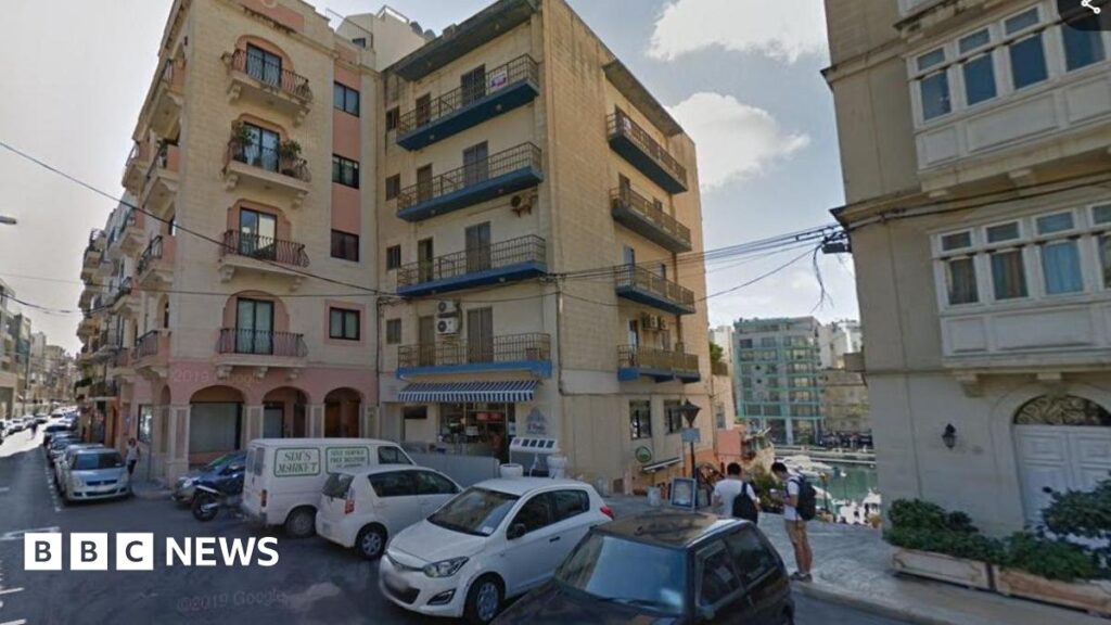 Gwynedd man, 25, dies in Malta balcony fall A road of hotels and tiled apartments overlooking the sea. Cars are parked outside. The bottom of one building has a blue and white striped awning and shop, and the other has pink arches and columns.