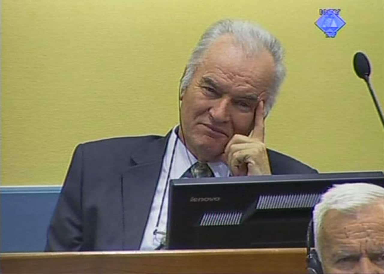 Middle aged man with white hair, wearing a suit, a finger pressed to his temple, a greenish yellow background, in a courtroom, with a microphone and screen visible.