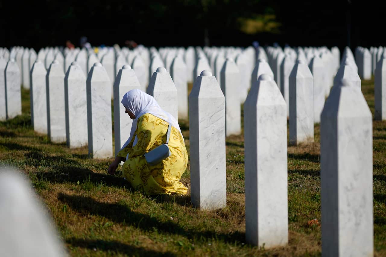 A woman wearing a white headscarf mourning next to the grave of her relative, in a field of identical white rectangular vertical tomb stones. 