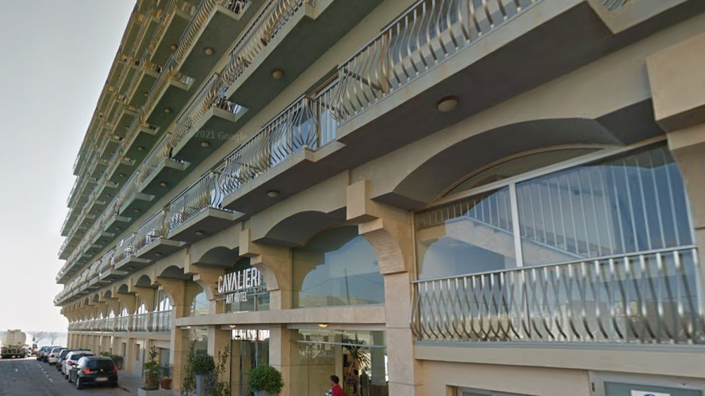 The outside of a sand-coloured hotel with a row of balconies above an entranceway which has lots of glass windows. In front are a row of cars parked along a road.