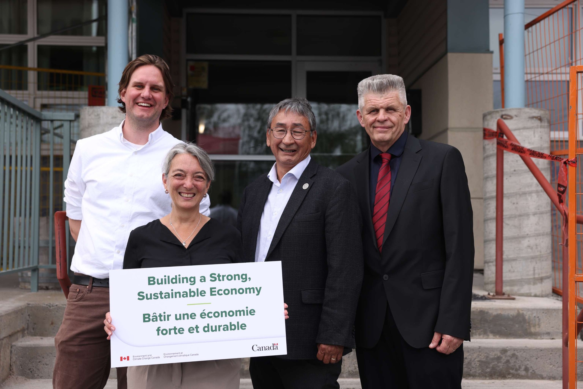 Four adults in business attire stand smiling in front of a building entrance. Minister Dabrusin is in front holding a bilingual sign with the Government of Canada logo and the Environment and Climate Change Canada logo. Text: Building a Strong, Sustainable Economy / Bâtir une économie forte et durable.