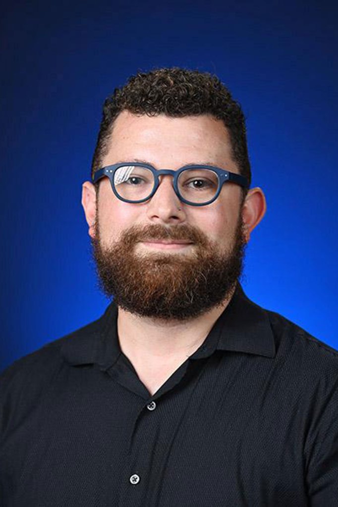 A man wearing blue glasses and a black shirt stands in front of a blue background