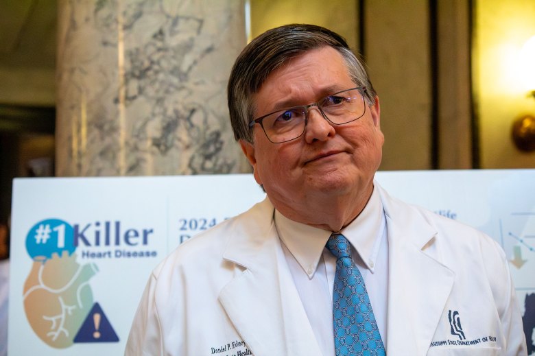 An older man in a doctor's coat looks off to the right while listening to something. A sign behind him read #1 Killer Heart Disease