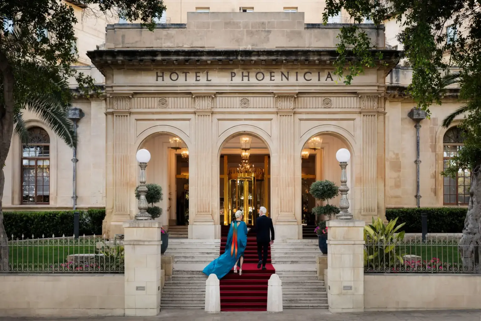 Elegant entrance of The Phoenicia Malta with two guests walking up the red-carpeted stairs under grand arches, surrounded by classical architecture.