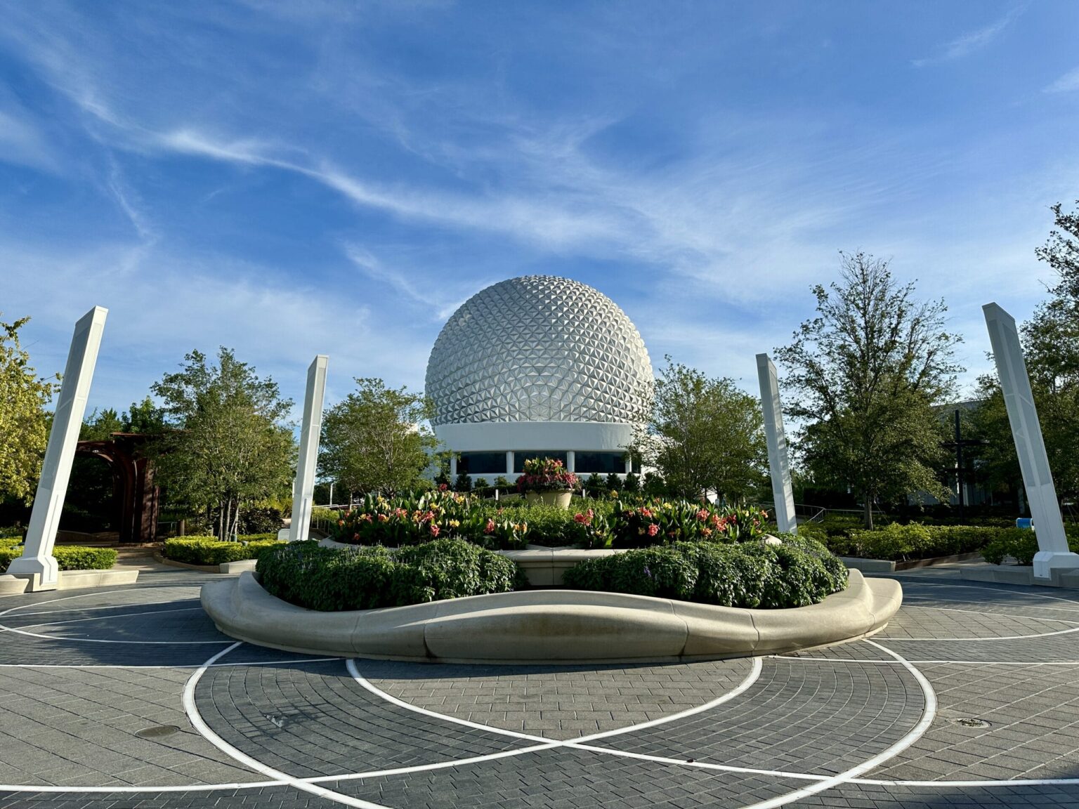 PHOTO REPORT: EPCOT 7/8/2025 (Pin Tuesday, Bobbing Heads in Norway, and a Dramatic Figment Moment) A large geodesic sphere at EPCOT stands behind a landscaped garden with flowers and trees under a blue sky.