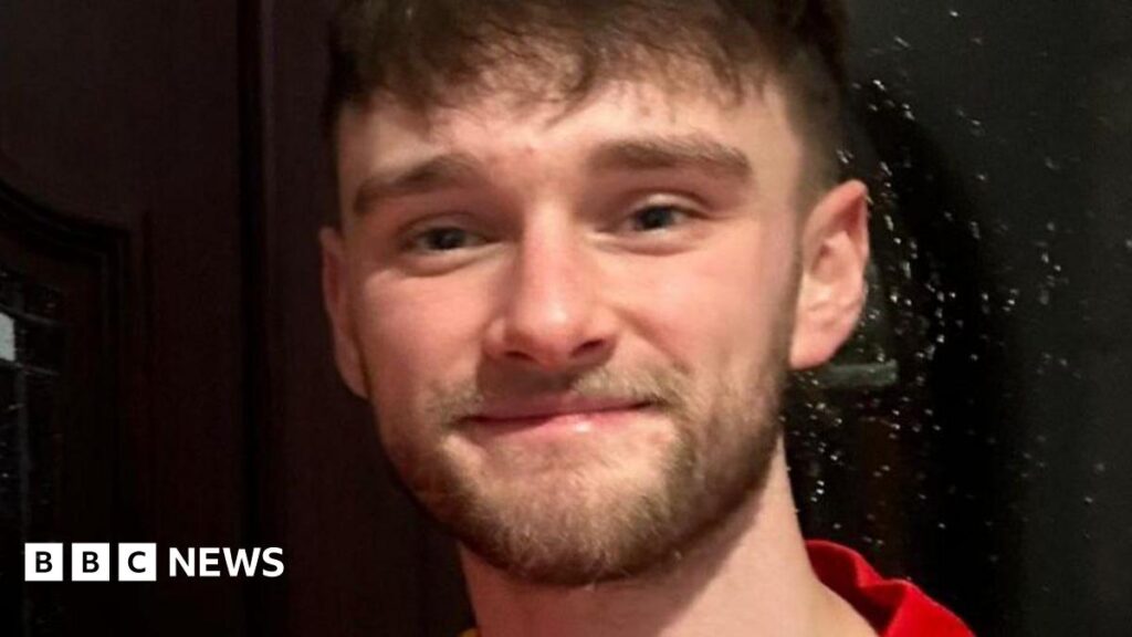 Kieran Thomas Hughes smiling into the camera. he has a short brown beard and brown hair with a fringe. He is wearing a red shirt.