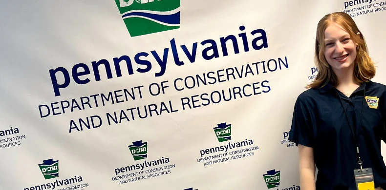 A person wearing a dark shirt and badge stands smiling in front of a backdrop with the Pennsylvania Department of Conservation and Natural Resources logo and text.