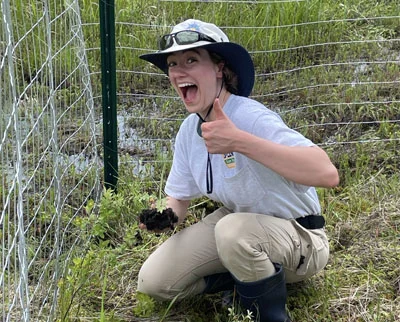 A smiling person in outdoor gear crouches by a wire fence, holding soil and giving a thumbs up, surrounded by green grass and plants.