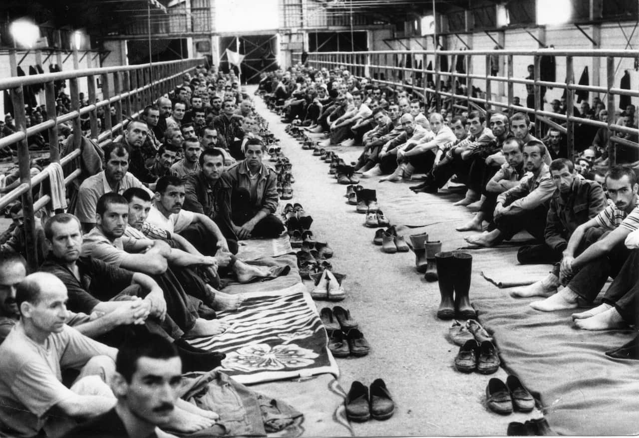 Dozens of men looking at the camera. Black and white, their shoes off and lined up in front of them.