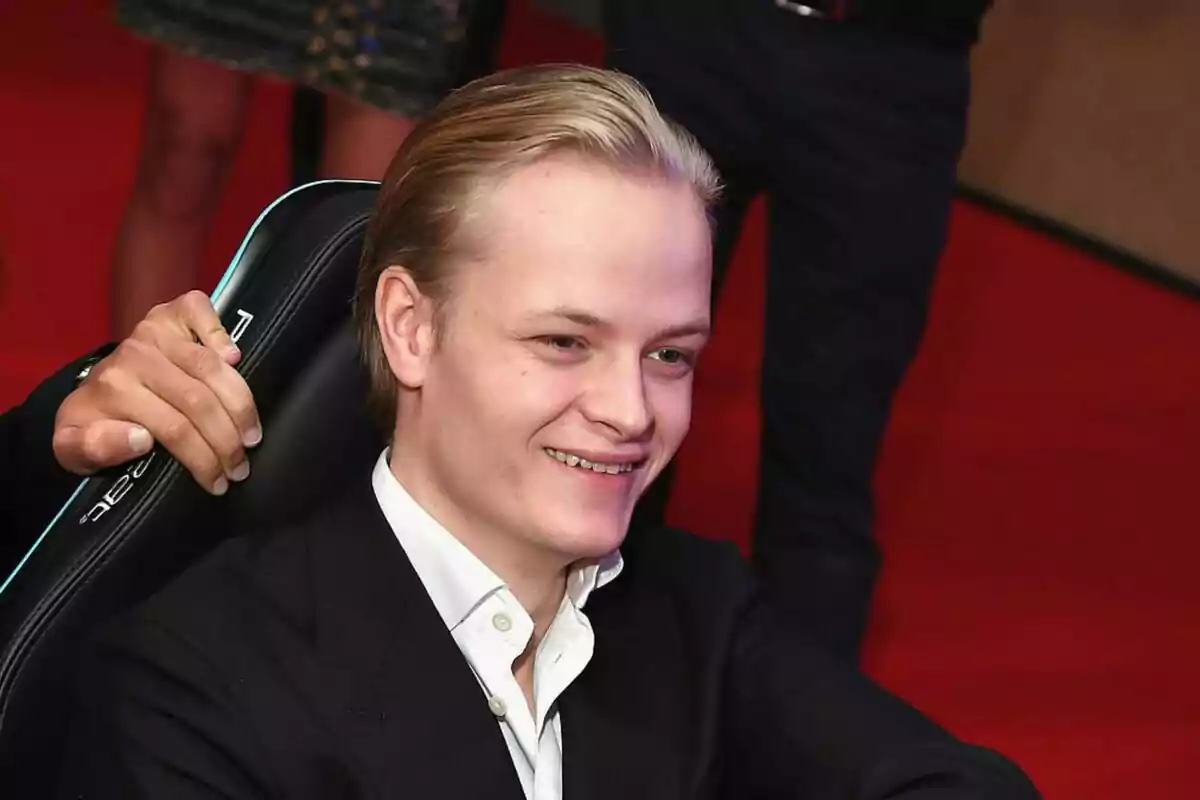Marius Borg slicked back, wearing a white shirt and dark jacket, smiling while sitting in a chair with a red background.